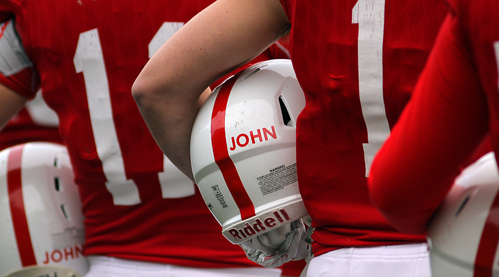 An unidentified St. John's player (shown from the back) stands with his helmet under his arm. The word JOHN is clearly visible, in red capital letters on a white helmet. (Photo by Wade Gardner, d3photography.com)
