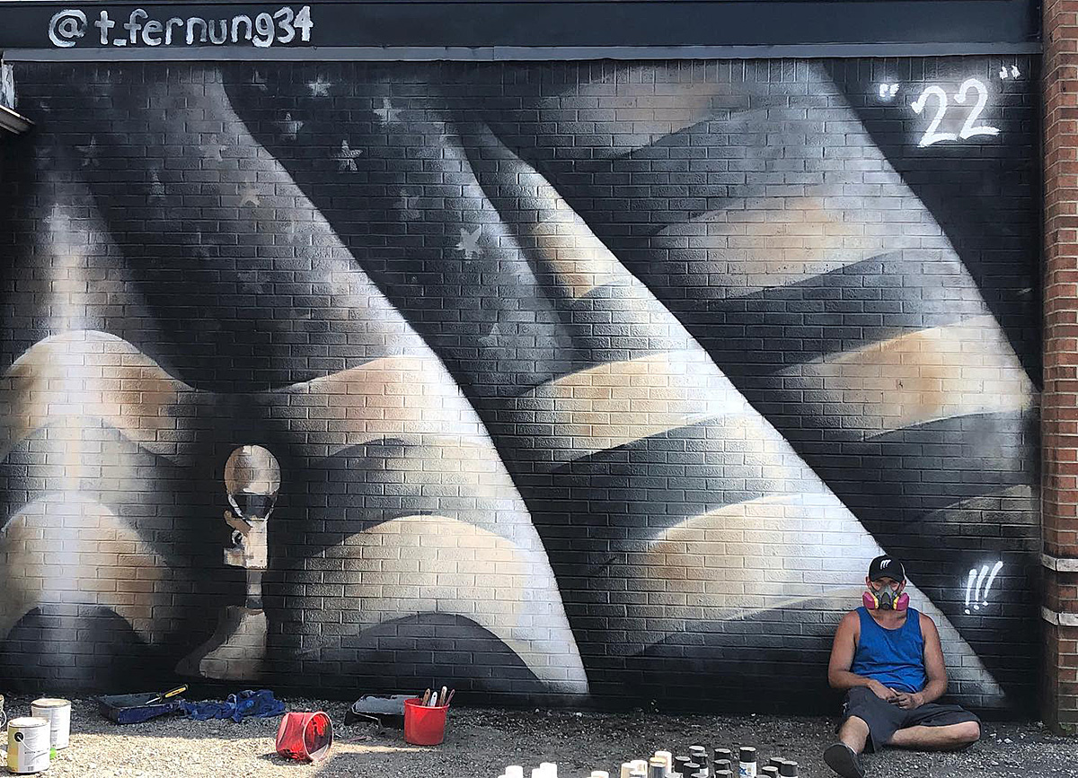 Tanner Fernung sits on the ground in front of a mural of his, an American flag spray-painted on a brick wall with the number 22 in white in the upper right-hand corner.